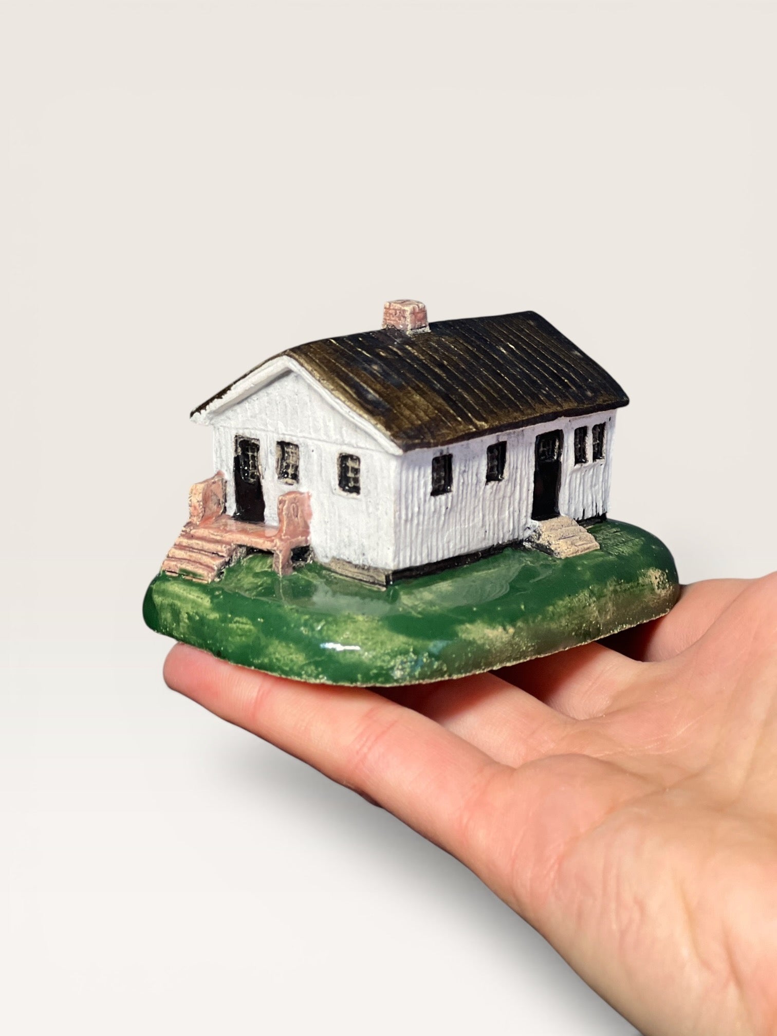 Ceramic miniature replica of a white cottage with wooden porch stairs and a dark roof held in a hand.